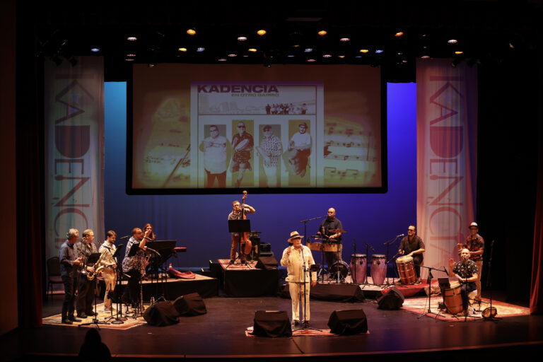 Kadencia Orchestra - The Cultural Current Large ensemble performing on a theater stage with a projection screen showing a poster and banners on both sides.