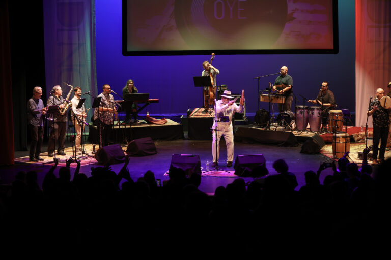 3M9A8290final - The Cultural Current Jazz band performing on a colorfully lit stage; center-front vocalist in white with a hat sings into a microphone while audience silhouettes raise their hands.