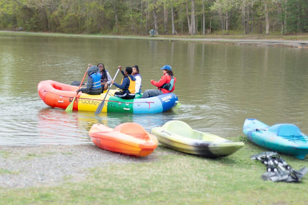 MOUNTAINFILM 2025-49_edited - The Cultural Current Group of people wearing life jackets paddling a large colorful inflatable raft on a calm lake with trees in the background.