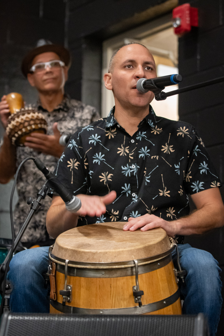 Maurice "Tito" Sanabria - The Cultural Current Man wearing a palm-tree shirt plays a conga drum and sings into a microphone, with another musician in the background holding an object and wearing glasses and a hat.