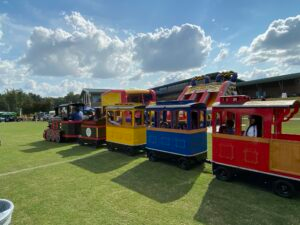 SoulfulSunday-2024-Kids-300x225 - The Cultural Current Colorful miniature train with red, yellow, and blue cars parked on a grassy field at an outdoor event, with a blue sky and clouds above.
